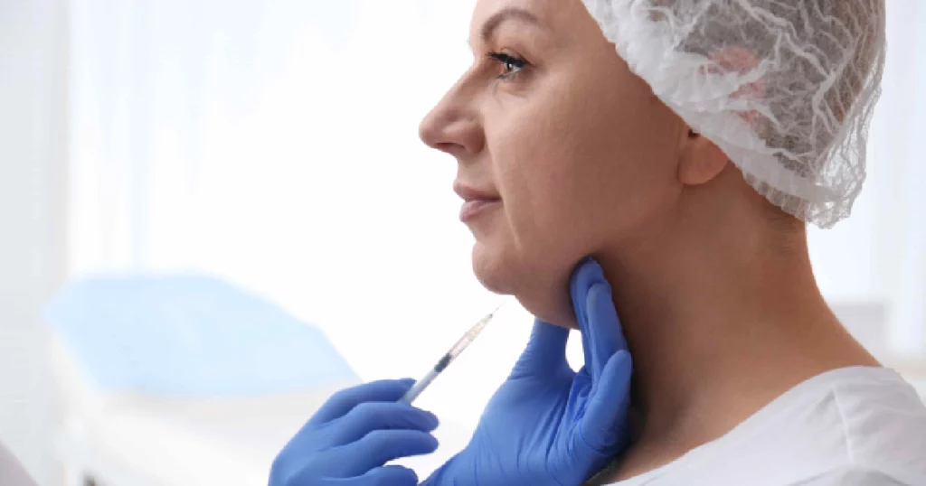 A woman wearing a hair cap receives a Kybella injection under her chin from a professional wearing blue gloves to reduce double chin fat in Draper, UT